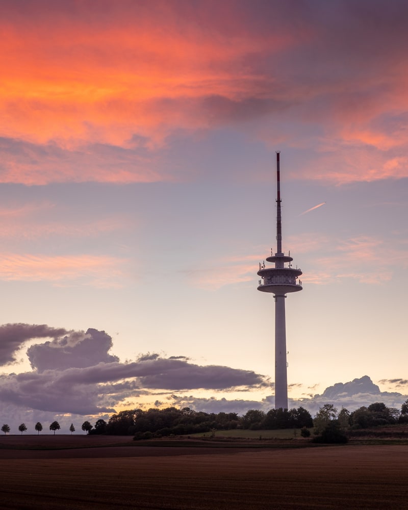 The Braunschweiger Fernmeldeturm on the Steinberg under orange glowing clouds as a plane passes in the background.