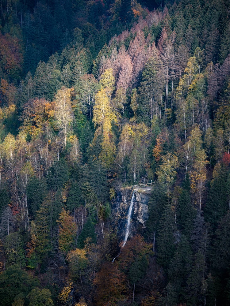 The "Romkerhaller Wasserfall" a waterfall is seen from above as the sun illuminates some fall colored trees behind it.