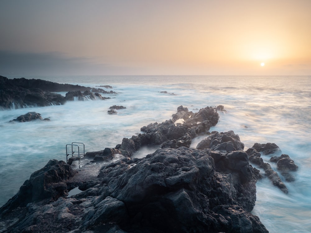 Long-exposure shot of a lava rock formation in the ocean with the setting sun in the background.